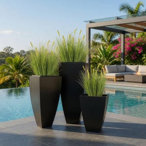 May include: Three black, geometric planters of varying sizes, each filled with tall, green ornamental grass. The planters are arranged on a tiled patio near a swimming pool, with a modern outdoor seating area in the background.