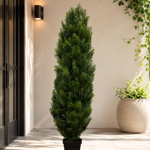 May include: A tall, slender artificial cedar topiary tree with dense, vibrant green foliage. The tree is shaped into a narrow, upright column and is potted in a simple, black cylindrical container, standing on a light-coloured patio floor.