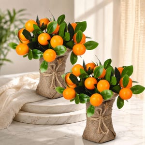 Pode incluir: Duas laranjeiras artificiais em vasos envoltos em juta. Cada planta apresenta frutos esféricos de cor laranja brilhante e folhas verde-escuras. Os vasos são amarrados com corda natural, criando um aspeto rústico para estes arranjos botânicos de mesa.
