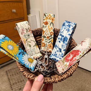 Puede incluir: Una colección de llaveros de tela para muñeca con estampados florales en una cesta tejida. Los llaveros presentan varios diseños, incluyendo girasoles, margaritas y rosas, con herrajes plateados. La paleta de colores incluye azul, amarillo, naranja y rojo.
