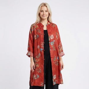 Peut inclure: Une veste mi-longue de couleur rouille à manches longues, ornée d'un imprimé floral aux tons bleu, vert et blanc. Le vêtement présente un col officier, une fermeture frontale avec des boutons de style brandebourg et des poignets larges.
