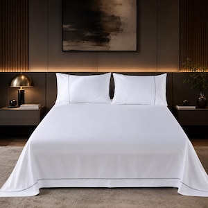 May include: A white bed sheet set featuring two matching pillowcases and a flat sheet, each adorned with a thin, dark gray embroidered border detail. The bedding is smooth and crisp, providing a clean, neutral aesthetic for a bed setup.