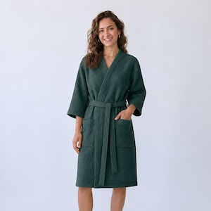 May include: A dark forest green robe featuring a textured waffle-knit pattern, a V-neckline, three-quarter length sleeves, two front patch pockets, and a matching fabric tie belt that secures at the waist for a structured and comfortable fit.