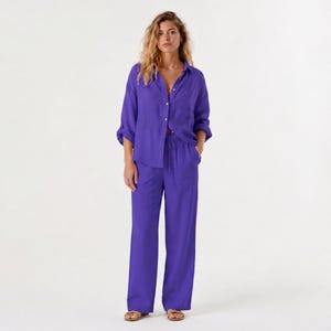 May include: A vibrant purple two-piece set consisting of a long-sleeved button-down shirt with a collar and matching wide-leg trousers. The trousers feature a drawstring waistband and a relaxed fit, while the shirt has a single chest pocket.