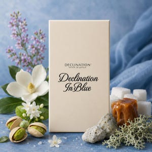 May include: A rectangular cream-colored perfume box with the text Declination extrait de parfum and Declination In Blue printed in black. The box is surrounded by white flowers, green pistachio nuts, a caramel cube, white sugar cubes, and lichen.