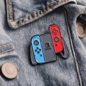Op de afbeelding: Een Switch-controller pin met blauwe en rode accenten, geplaatst op een spijkerjasje. De pin heeft een grijze console met een blauwe controller aan de linkerkant en een rode controller aan de rechterkant. De jas heeft zichtbare stiksels en een knoop.