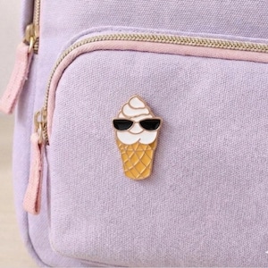 May include: A gold-colored ice cream cone pin with a white swirl of ice cream and black sunglasses. The pin is attached to a light purple backpack with pink zippers and straps. The ice cream cone pin is a fun accessory.
