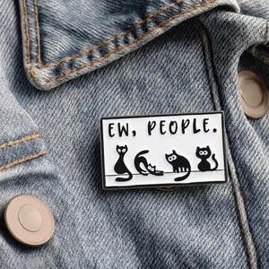 Könnte beinhalten: Rechteckiger Emaille-Anstecker mit schwarzem Rand und der Aufschrift "EW, PEOPLE." Der Anstecker zeigt vier schwarze Katzen-Silhouetten in verschiedenen Posen. Der Anstecker ist an einer Jeansjacke befestigt.