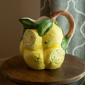 May include: A ceramic pitcher with a lemon design. The pitcher is yellow with brown and green accents, featuring a handle and a spout. The pitcher is decorated with lemon slices and leaves. It is sitting on a round wooden table.