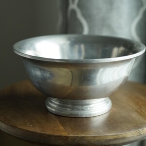 May include: A silver metal bowl with a rounded shape and a pedestal base. The bowl is sitting on a round wooden table. The bowl has a shiny, reflective surface.