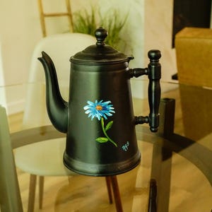 May include: A matte black metal teapot featuring a hand-painted blue daisy with a green stem and leaves on the front. The teapot has a curved spout, a lid with a small knob handle, and a vertical black handle with decorative turned detailing.