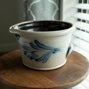 May include: A cream-colored ceramic bowl with a dark brown interior and a blue floral design. The bowl has a handle and a spout. It sits on a round wooden table. The background includes a window with blinds.