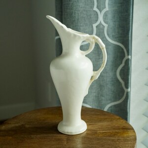 May include: A tall, off-white ceramic pitcher featuring a slender neck, a flared pouring spout, and a curved handle. The vessel has a smooth, matte finish with a rounded body that tapers down to a circular base, presenting a clean, solid-colored design.