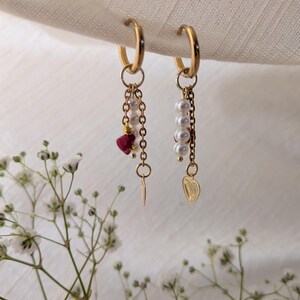 Peut inclure: Une paire de boucles d'oreilles créoles dorées avec des breloques pendantes. Chaque boucle comporte une chaîne dorée, des perles blanches et un petit cœur doré. L'une des boucles inclut une perle en pierre rouge irrégulière.
