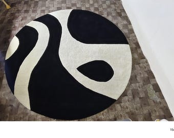 Puede incluir: Una alfombra circular con un patrón abstracto de alto contraste de formas orgánicas fluidas en negro sólido y blanco roto. El diseño consta de secciones curvas entrelazadas que crean un efecto visual gráfico audaz en toda la superficie.