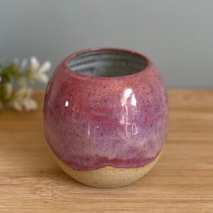 May include: A small, round ceramic vase with a textured, glossy finish. The top half is a vibrant pink-purple, transitioning to a beige base. The interior is a light blue-grey. The vase sits on a wooden surface.