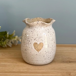 May include: A speckled ceramic vase with a heart design. The off-white vase has a ruffled rim and is covered in small black speckles. The heart is a lighter tan colour. The vase is on a wooden surface.