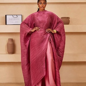 May include: A long, flowing, pleated magenta dress with a high neckline and wide sleeves. The dress is cinched at the waist and has a split skirt revealing a lighter pink underlayer. A shopping bag with the text "SHESABI" is visible.
