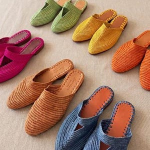 Puede incluir: Una colección de mules de rafia trenzada en varios colores, incluyendo verde lima, amarillo, naranja, beige, azul y magenta, junto con varios cinturones trenzados a juego con hebillas rectangulares y un cinturón azul enrollado.