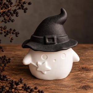 Può includere: Una statuetta in ceramica bianca a forma di fantasma arrotondato che indossa un cappello da strega nero con una fibbia. Il fantasma ha due occhi circolari, una piccola bocca rotonda e una bacchetta a forma di stella in un braccio.