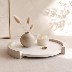 Può includere: Un vassoio decorativo dai toni neutri con un design rotondo, con un piccolo vaso con fiori secchi, una candela accesa in un portacandele di vetro e un anello d'oro. Il vassoio ha un bordo rialzato e un'estetica minimalista.