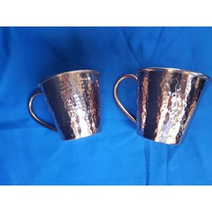 May include: Two copper-coloured mugs with hammered textures and curved handles. The mugs have a shiny finish and are set against a blue background. The mugs are designed for beverages.