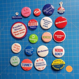 May include: A selection of vintage political campaign badges and pins, featuring various candidates and slogans. The badges are round and oval, with text and graphics in red, white, blue, and green. Some badges include the names Nixon, McGovern, and Ford.