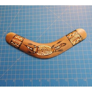 May include: A wooden boomerang with a curved shape, featuring a hand-painted design. The boomerang has a brown base color with decorative patterns in shades of brown, white, and black. A central illustration depicts a stylized crocodile.