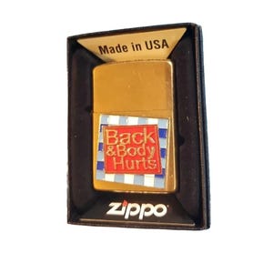 May include: A gold-colored Zippo lighter with the text "Back & Body Hurts" on a red and blue checkered background. The lighter is in a black box with "Made in USA" printed on the top.