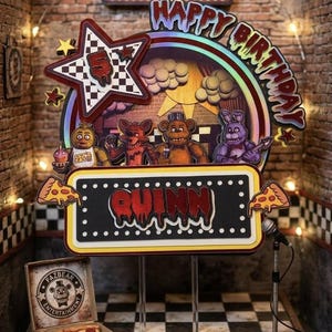 May include: A colorful birthday cake topper featuring characters from the video game Five Nights at Freddy's. The design includes a star, a sign that says "Quinn", and the words "Happy Birthday". The scene is set in a brick-walled room.