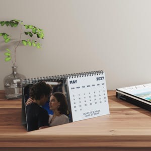 May include: A spiral-bound desk calendar for May 2027 featuring a photograph of a couple facing each other. The calendar displays the month, year, and days of the week, with the text "HEART OF A HAWK HOME AT BRIAR" printed below the dates.