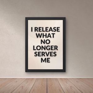 May include: A framed print with the words "I RELEASE WHAT NO LONGER SERVES ME" in black. The print has a cream-coloured background and is in a black frame. It's displayed on a wall above a wooden floor, creating a simple, modern aesthetic.