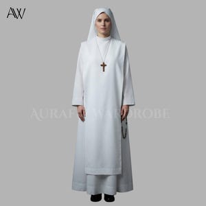 Może przedstawiać: Biały habit o pełnej długości, składający się z tuniki z długimi rękawami, szkaplerza i pasującego nakrycia głowy. Na szyi znajduje się drewniany krzyż na łańcuszku, a przy talii wisi czarny różaniec. Odzież jest w jednolitym białym kolorze.