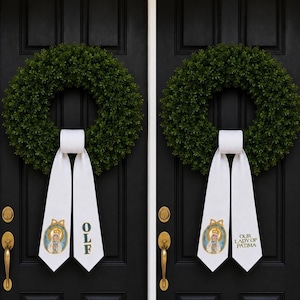 May include: Two dark green boxwood wreaths hanging on black doors. Each wreath features a white fabric ribbon streamer. One ribbon displays the text OLF and an image of Our Lady of Fatima, while the other displays the text Our Lady of Fatima.