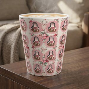 May include: A tall, white ceramic coffee mug with a floral and portrait pattern. The design features oval portraits of a woman in a nun's habit, surrounded by pink roses. The mug is filled with coffee, and steam rises from the top.