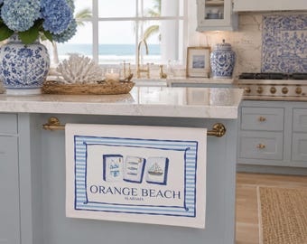 May include: A white rectangular kitchen towel featuring a blue striped border and a central graphic of three mahjong tiles displaying a bikini, three fish, and a sailboat. The text reads ORANGE BEACH ALABAMA in a dark blue serif font.