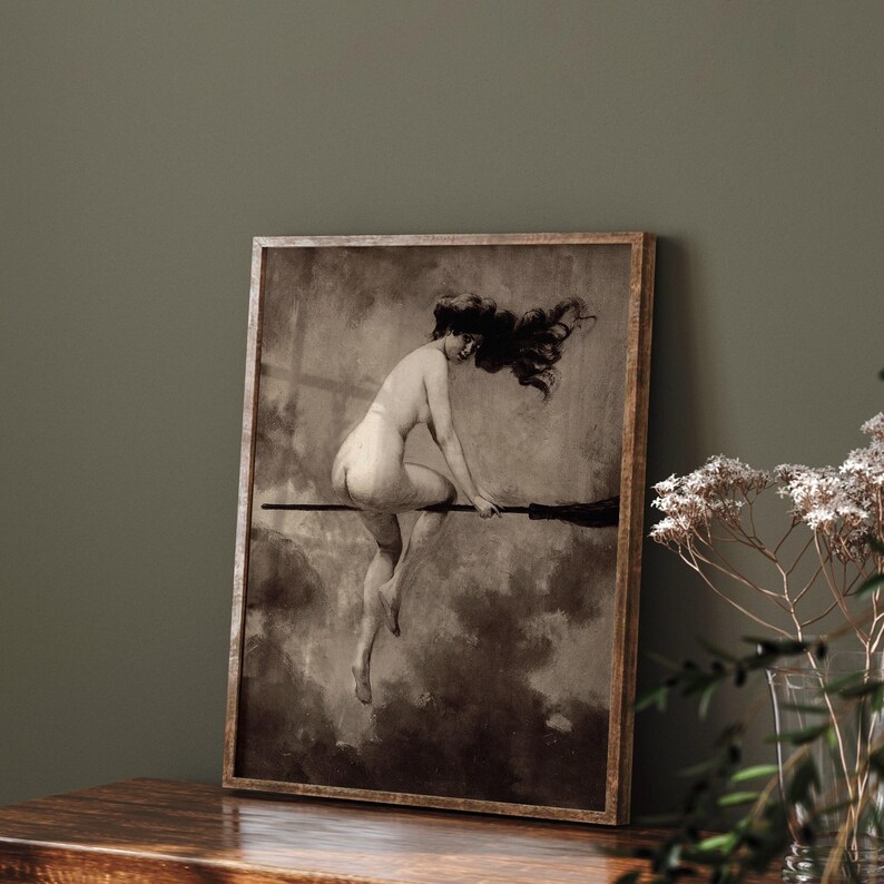 May include: A framed sepia-toned art print depicting a figure with long flowing hair riding a broomstick through a cloudy sky. The figure is shown in profile, positioned horizontally across the broom, set within a simple wooden frame.