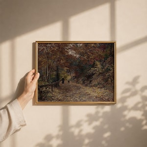 Puede incluir: Un cuadro de paisaje rectangular en un marco dorado que representa un camino a trav&eacute;s de un bosque en oto&ntilde;o. La escena muestra hojas amarillas ca&iacute;das en el suelo, &aacute;rboles densos con follaje marr&oacute;n y naranja, y una figura peque&ntilde;a con un perro.