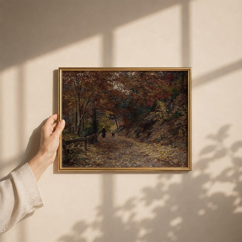 Puede incluir: Un cuadro de paisaje rectangular en un marco dorado que representa un camino a trav&eacute;s de un bosque en oto&ntilde;o. La escena muestra hojas amarillas ca&iacute;das en el suelo, &aacute;rboles densos con follaje marr&oacute;n y naranja, y una figura peque&ntilde;a con un perro.