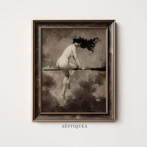 May include: A framed wall art print featuring a sepia-toned illustration of a nude figure riding a broomstick against a cloudy sky. The artwork is enclosed in a dark, distressed wooden frame with gold accents. The text AESTIQUEA is printed below.