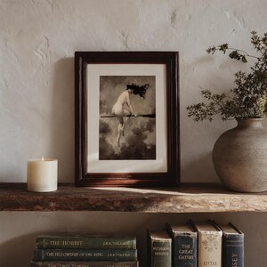 May include: A framed art print featuring a monochromatic illustration of a figure riding a broomstick against a cloudy sky. The dark wood frame surrounds a white mat board. The artwork depicts a person in motion with flowing hair.