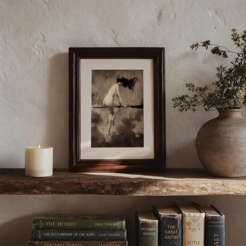 May include: A framed art print featuring a monochromatic illustration of a figure riding a broomstick against a cloudy sky. The dark wood frame surrounds a white mat board. The artwork depicts a person in motion with flowing hair.