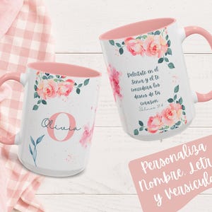 May include: Two white ceramic mugs with pink interiors and handles. Floral designs with pink roses and green leaves decorate the mugs. One mug has the name "Olivia" and a letter "O". The other mug has Spanish text. The mugs are on a pink and white checkered background.