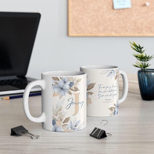 May include: Two white ceramic mugs with floral designs. One mug features a large letter "J" and the name "Jenny" with blue and tan flowers. The other mug has text in Spanish. Both mugs have a handle.