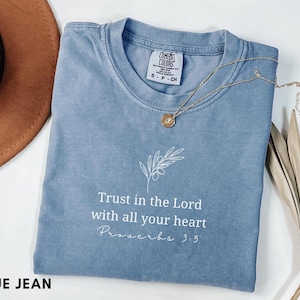 Puede incluir: Una camiseta de manga corta color azul jean con una ilustración de líneas blancas de una rama de olivo sobre el texto "Trust in the Lord with all your heart Proverbs 3:5". La camiseta tiene una etiqueta de la marca Comfort Colors.