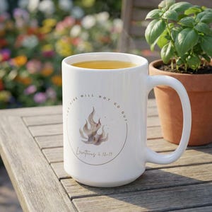 May include: White ceramic mug with a large handle, filled with a light yellow liquid. The mug features a circular design with a flame illustration and the text "THE FIRE WILL NOT GO OUT" and "Leviticus 6:12-13". The mug is on a wooden table.