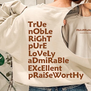 May include: A beige crewneck sweatshirt featuring brown text on the back that reads TrUe, nObLe, RiGhT, pUrE, LoVeLy, aDmIrAbLe, EXcEllent, pRaiseWortHy. The front chest area displays the text PhiLiPPiaNs 4:8 and THINK ON THESE THINGS.