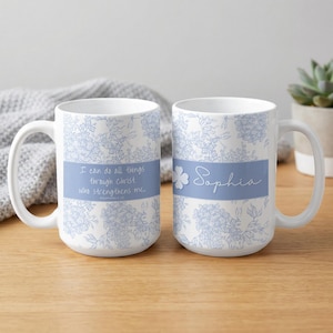 Op de afbeelding: Twee witte keramische mokken met een blauw bloemenpatroon. Eén mok heeft de tekst "I can do all things through Christ who strengthens me". De andere mok heeft de naam "Sophia".