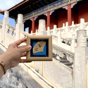 Puede incluir: Una pequeña obra de arte cuadrada con marco de madera que presenta una hoja de ginkgo preservada con caligrafía china sobre un fondo azul. La obra de arte se sostiene frente a un edificio chino tradicional.