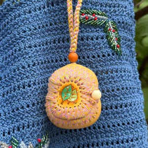 May include: A blue crocheted bag featuring a round, tan, carved wooden pendant suspended by a braided yellow and pink cord. The pendant is decorated with pink and yellow stitching and a central illustration of two green leaves with a small white bead.